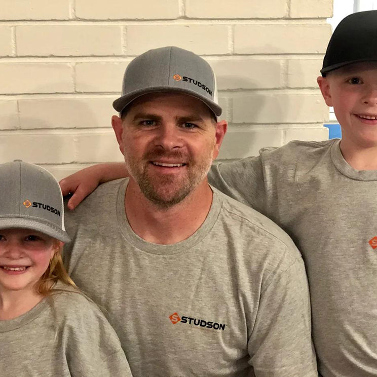 Man and two children wearing gray 'Studson' hats and shirts against a light brick wall.