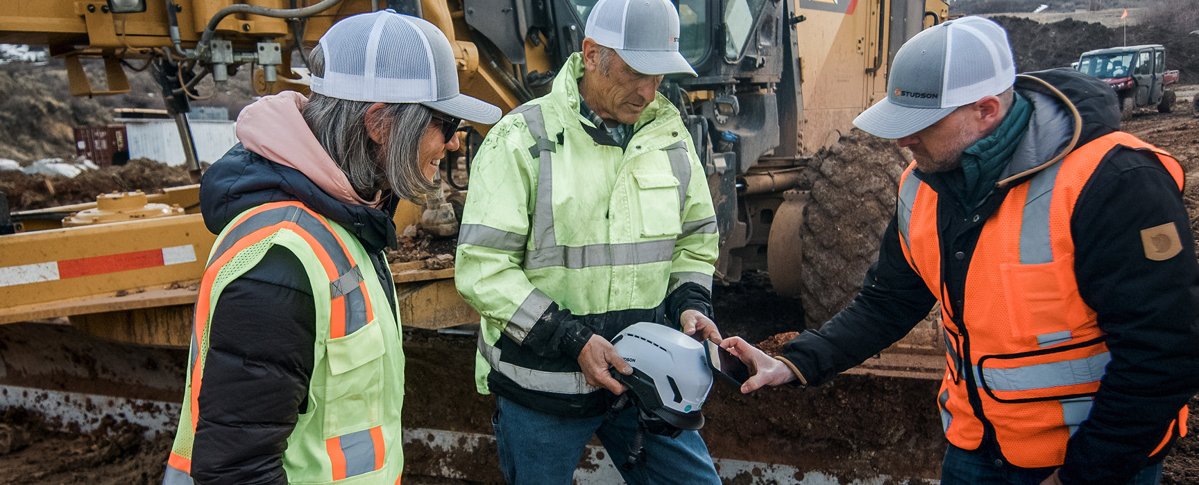 Three construction workers in safety gear discussing near heavy machinery on a job site.