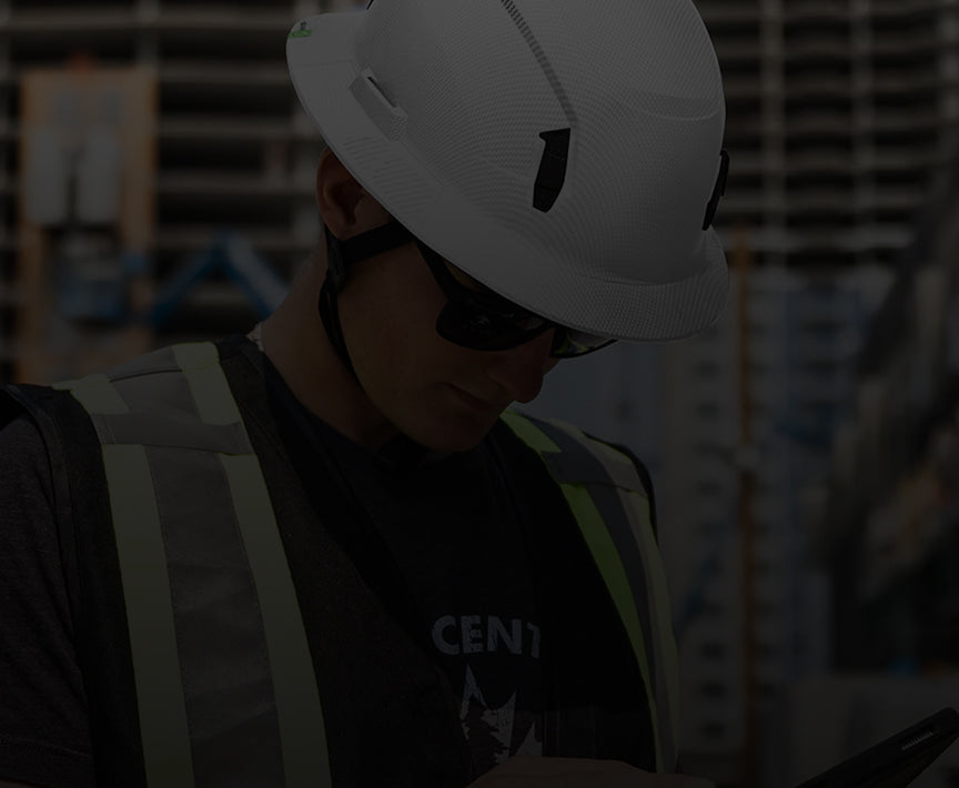 Person wearing a white helmet and sunglasses, looking at a device in an industrial setting.
